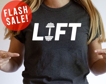 May include: A dark gray t-shirt with the word "LIFT" printed in white with a dumbbell graphic in the middle of the "I". The model is wearing the shirt and pointing to it. The text "FLASH SALE!" is in a red circle in the top left corner of the image. The text "MORE SHIRT COLORS" is in a red rectangle in the bottom right corner of the image.