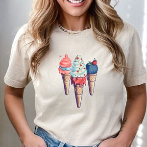 May include: A white t-shirt with a graphic of three ice cream cones. The ice cream cones are topped with different flavors of ice cream, including pink, blue, and purple. The ice cream cones are on a white background.