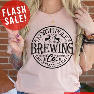 May include: A light pink t-shirt with a circular black graphic that reads "North Pole Brewing Co. Christmas Spirits." The shirt has the text "Flash Sale!" in a red circle and "More Shirt Colors" in a red rectangle.