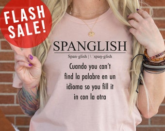 Op de afbeelding: Een roze T-shirt met het woord "Spanglish" gedefinieerd als "Wanneer je het woord niet kunt vinden in één taal, vul je het dan aan met de andere taal". Het shirt heeft ook de tekst "Meer shirtkleuren" in rood.