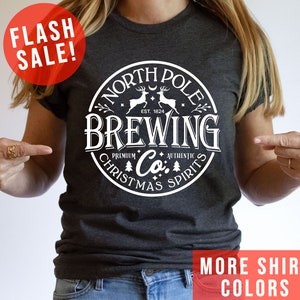May include: Dark gray t-shirt with a white circular graphic that reads "North Pole Brewing Co. Premium Authentic Christmas Spirits." The shirt also has the text "Flash Sale!" and "More Shirt Colors" in red.