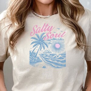 May include: A beige t-shirt featuring a graphic of a palm tree, sun, and waves in blue and pink. The text "Salty Soul" is written in pink cursive.