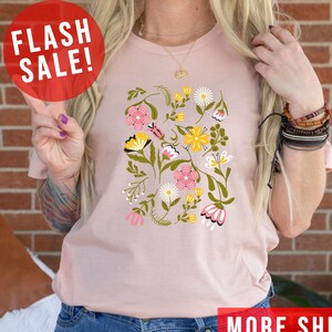 May include: A pink t-shirt with a floral print featuring pink, yellow, and white flowers. The shirt has a crew neck and short sleeves. The text "MORE SHIRT COLORS" is visible in the bottom right corner of the image.