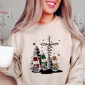 May include: Beige sweatshirt featuring a Christmas design with three snowmen wearing hats and scarves. The snowmen have the words "Faith, Hope, Love" below them. A cross and trees are also part of the design. The brand "DazzleTees" is printed on the top left.