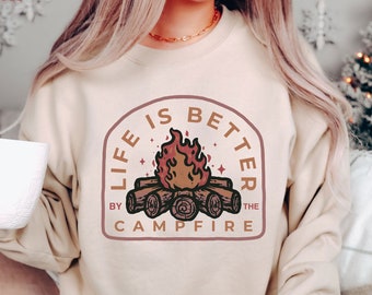 Könnte beinhalten: Ein beiger Pullover mit einem Grafikdruck eines Lagerfeuers mit dem Text "Life is better by the campfire".