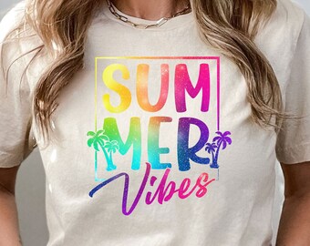 May include: A beige t-shirt with the words "SUMMER VIBES" in a colorful, rainbow-gradient design. The text is framed by a pink square and includes palm tree graphics. The shirt is short-sleeved.