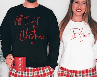 May include: A black sweatshirt with the text "All I want for Christmas..." in red, and red plaid pyjama bottoms. A white sweatshirt with the text "... Is You" in red, and red plaid pyjama bottoms. Both are holding mugs.