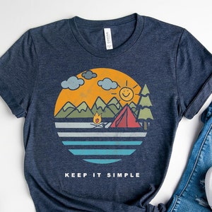 May include: A navy blue t-shirt with a retro-style illustration of a campsite with mountains, a lake, a campfire, and a tent. The text "Keep it simple" is printed below the illustration.