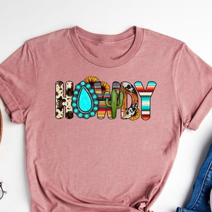 May include: A heather mauve t-shirt with the word "Howdy" printed on the front. The letters are decorated with various southwestern patterns, including turquoise, sunflowers, and cacti.
