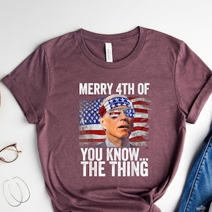 May include: Heather maroon t-shirt with the text "MERRY 4TH OF YOU KNOW... THE THING" and an image of a person wearing sunglasses and a headband with the American flag design. The shirt is paired with blue jeans and white sneakers.