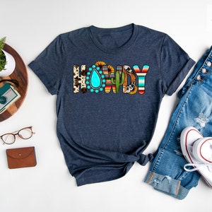 May include: A heather navy blue t-shirt with the word "Howdy" in colorful, patterned letters. The letters are decorated with southwestern designs, including sunflowers, turquoise, cacti, and cowhide.