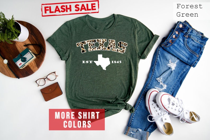 May include: A forest green t-shirt featuring a white outline of the state of Texas and the words "TEXAS EST 1845" in leopard print. The shirt is laid flat on a white background with a pair of blue jeans, white trainers, and other accessories.
