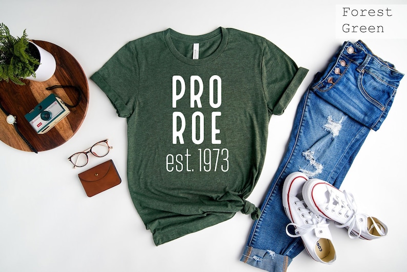 Op de afbeelding: Bosgroen T-shirt met witte tekst die "PRO ROE est. 1973" luidt. Het shirt ligt plat op een wit oppervlak met een spijkerbroek, een paar witte sneakers en een bruine leren portemonnee.
