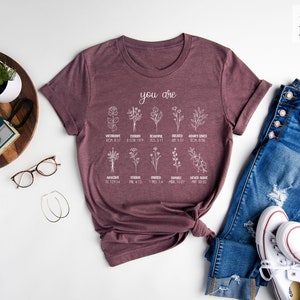 May include: A heather maroon t-shirt with a white graphic that says "you are" and lists different flowers with bible verses. The shirt is folded and laid on a white surface with a pair of glasses, a camera, a plant, and a pair of jeans and white sneakers.