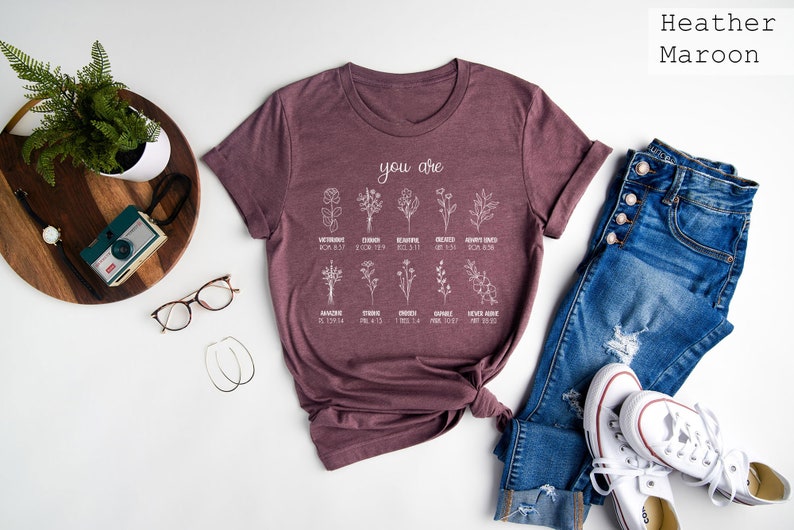 May include: A heather maroon t-shirt with a white graphic that says "you are" and lists different flowers with bible verses. The shirt is folded and laid on a white surface with a pair of glasses, a camera, a plant, and a pair of jeans and white sneakers.
