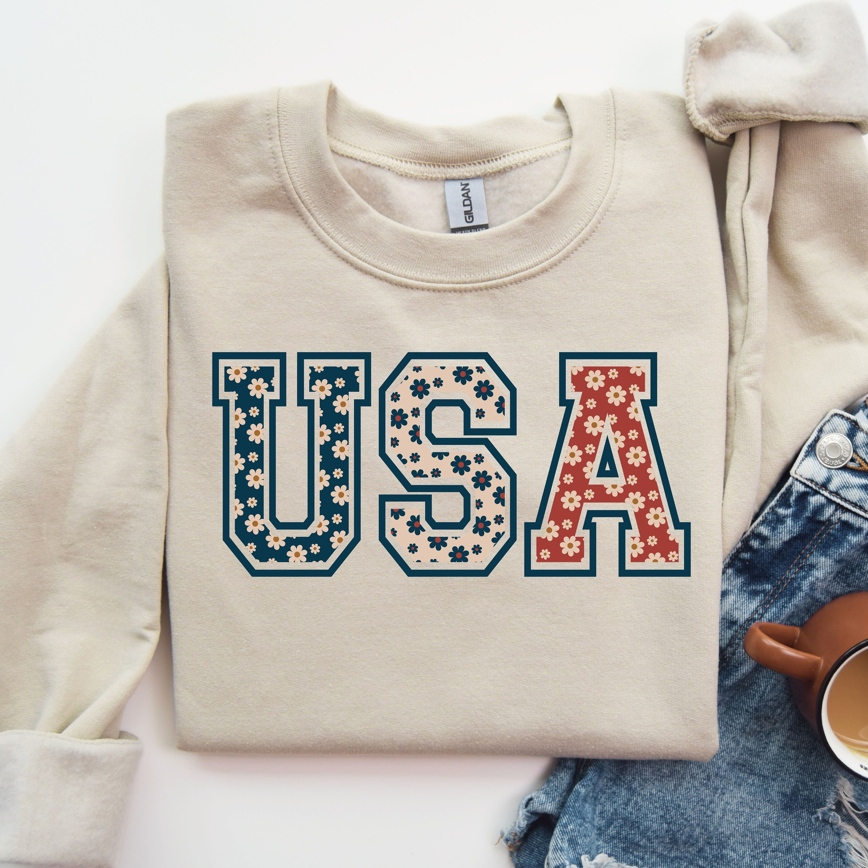 以下が含まれることがあります： ベージュ色のスウェットシャツ。文字「USA」はカレッジ風フォントで、ネイビーブルーの縁取りと赤、白、青の花柄で埋められており、愛国的なテーマを表現しています。