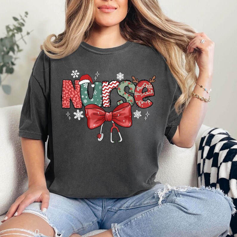 May include: A dark gray t-shirt with a Christmas-themed design that says "Nurse" in colorful letters with a red bow and a stethoscope. The letters are decorated with snowflakes and other festive elements.