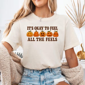 May include: Cream-colored t-shirt with the text "IT'S OKAY TO FEEL ALL THE FEELS" above a row of five orange jack-o'-lanterns with different facial expressions. The shirt is made of a soft material and is suitable for casual wear.