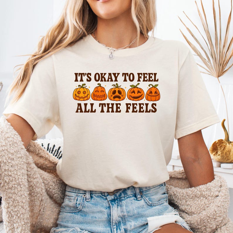 May include: Cream-colored t-shirt with the text "IT'S OKAY TO FEEL ALL THE FEELS" above a row of five orange jack-o'-lanterns with different facial expressions. The shirt is made of a soft material and is suitable for casual wear.