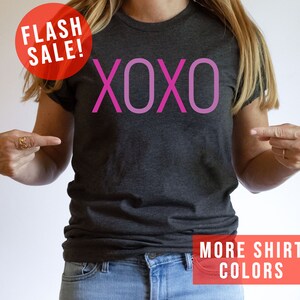 May include: A grey t-shirt with the word "XOXO" printed in pink on the front. The text is in a gradient of pink shades. The shirt is being modelled by a person. The image also includes the text "FLASH SALE!" and "MORE SHIRT COLORS".