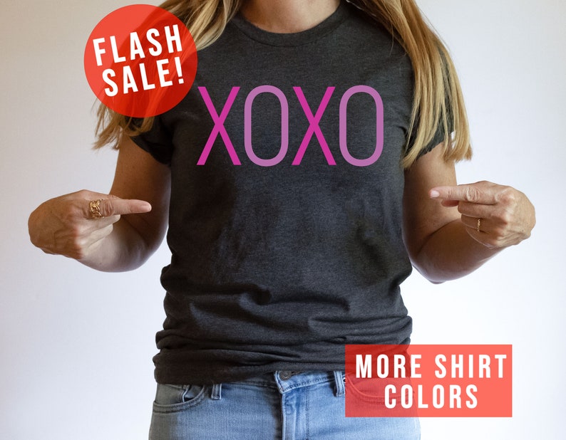 May include: A grey t-shirt with the word "XOXO" printed in pink on the front. The text is in a gradient of pink shades. The shirt is being modelled by a person. The image also includes the text "FLASH SALE!" and "MORE SHIRT COLORS".