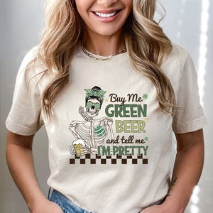 May include: A beige t-shirt with a graphic of a skeleton wearing sunglasses and a green headband holding a beer. The text on the shirt reads "Buy Me Green Beer and tell me I'm Pretty".