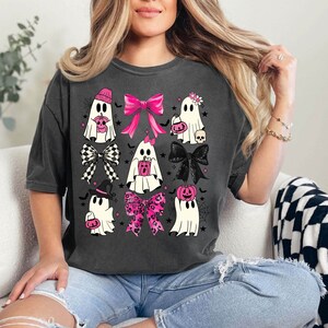 Puede incluir: Camiseta gris oscuro con una cuadr&iacute;cula de fantasmas de dibujos animados, cada uno adornado con lazos rosas, sombreros y detalles de calabaza. Los fantasmas est&aacute;n decorados con lazos, sombreros y accesorios tem&aacute;ticos de Halloween. Un dise&ntilde;o divertido y festivo para Halloween.