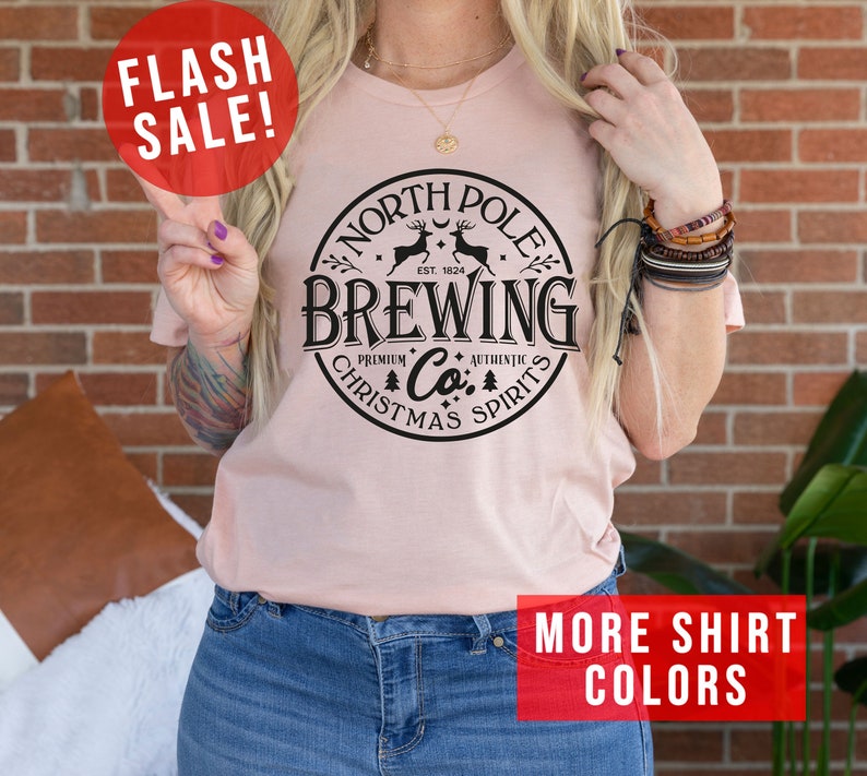 May include: A light pink t-shirt with a circular black graphic that reads "North Pole Brewing Co. Christmas Spirits." The shirt has the text "Flash Sale!" in a red circle and "More Shirt Colors" in a red rectangle.