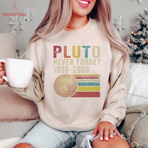 Op de afbeelding: Een beige sweatshirt met een vintage-stijl grafische afbeelding van Pluto, de tekst "Pluto Never Forget 1930-2006" en een gestileerde afbeelding van de planeet.