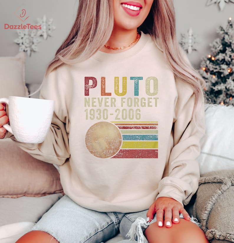 Op de afbeelding: Een beige sweatshirt met een vintage-stijl grafische afbeelding van Pluto, de tekst "Pluto Never Forget 1930-2006" en een gestileerde afbeelding van de planeet.