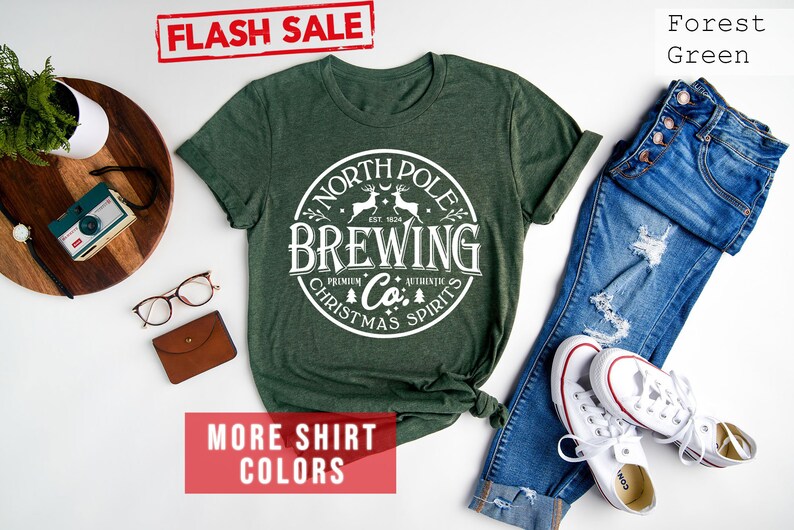 May include: Forest green t-shirt with a white circular graphic that reads "North Pole Brewing Co. Christmas Spirits." The shirt is paired with blue jeans and white sneakers. A "Flash Sale" banner is in the top left corner.