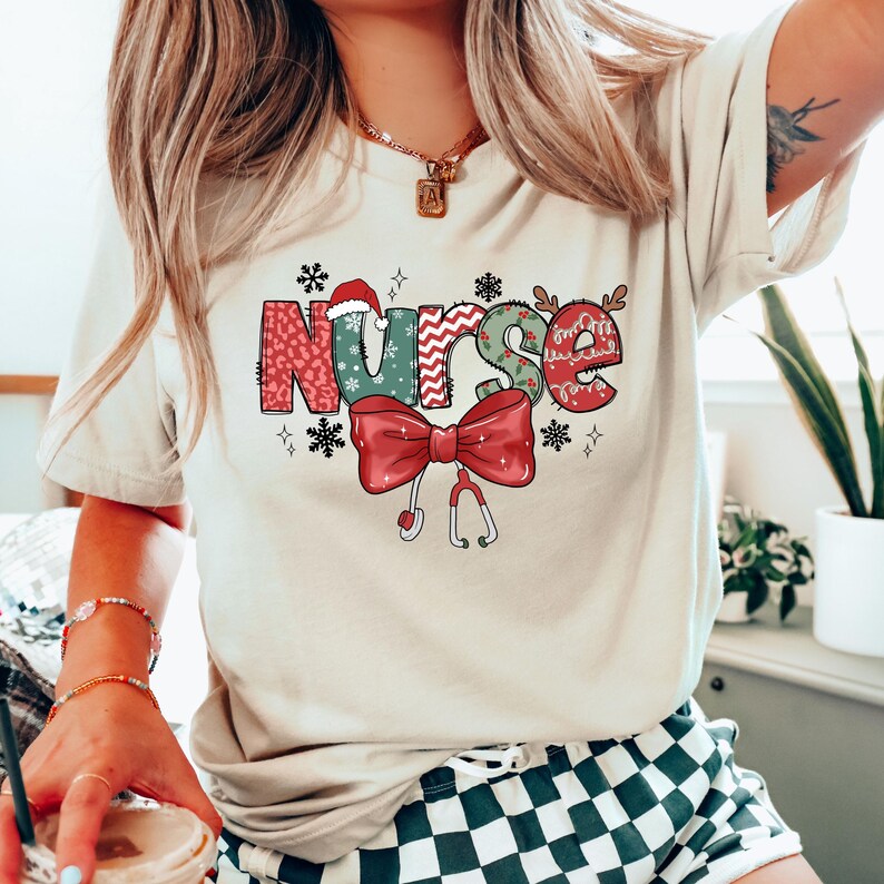 May include: A beige t-shirt with a festive Christmas-themed design featuring the word "NURSE" in colorful patchwork letters, a red bow with a stethoscope, and a Santa hat.