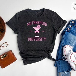 May include: A dark gray t-shirt with the text "MOTHERHOOD UNIVERSITY" in pink lettering. The text "EST. 1989" is also in pink lettering. A pink cocktail glass with cherries is in the center of the shirt.