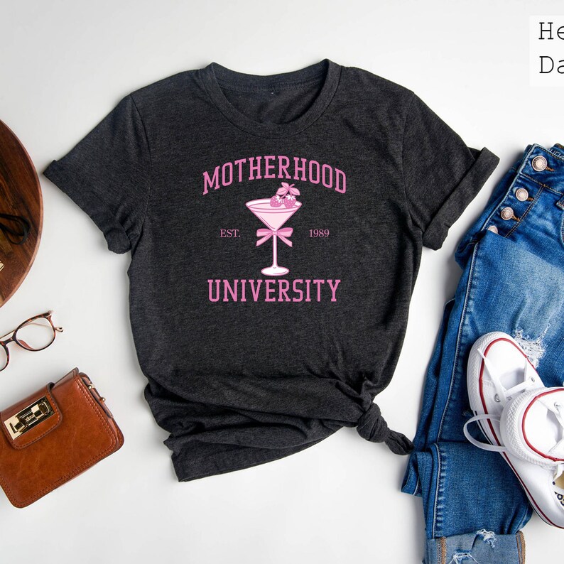 May include: A dark gray t-shirt with the text "MOTHERHOOD UNIVERSITY" in pink lettering. The text "EST. 1989" is also in pink lettering. A pink cocktail glass with cherries is in the center of the shirt.