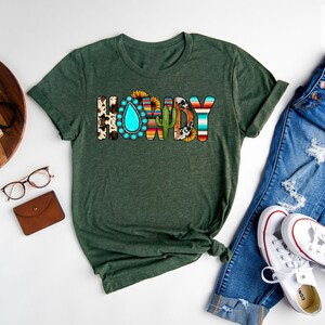 May include: Forest green t-shirt with the word "Howdy" in colorful letters with a southwestern design. The letters are decorated with a turquoise teardrop, a cactus, a sunflower, and a cowhide pattern.
