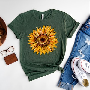 May include: A forest green t-shirt with a yellow sunflower graphic. The shirt is shown with blue jeans and white sneakers. The text "Forest Green" is visible in the top right corner of the image.
