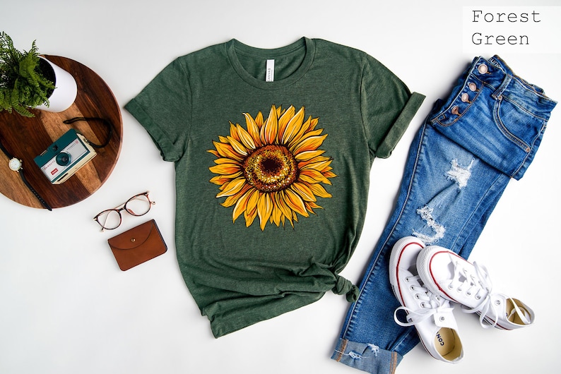 May include: A forest green t-shirt with a yellow sunflower graphic. The shirt is shown with blue jeans and white sneakers. The text "Forest Green" is visible in the top right corner of the image.