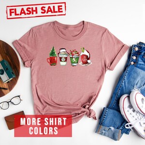 May include: Heather mauve t-shirt with a festive graphic of four holiday-themed beverages. The shirt features a Flash Sale banner and text that reads "MORE SHIRT COLORS". A pair of blue jeans and white sneakers are next to the shirt.
