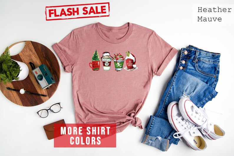 May include: Heather mauve t-shirt with a festive graphic of four holiday-themed beverages. The shirt features a Flash Sale banner and text that reads "MORE SHIRT COLORS". A pair of blue jeans and white sneakers are next to the shirt.
