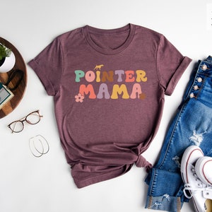 May include: A heather maroon t-shirt with the text "POINTER MAMA" in colourful retro-style lettering. The shirt is shown laid out on a white surface with a pair of jeans, white trainers, and other accessories.