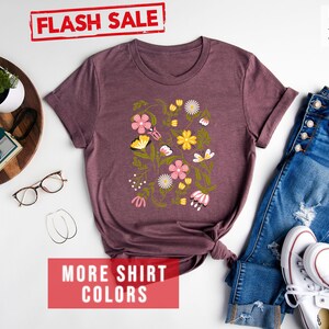 May include: A heather maroon t-shirt with a floral print featuring pink, yellow, and green flowers and butterflies. The shirt is displayed on a white background with a pair of jeans, white sneakers, and a wooden tray with a camera and glasses.