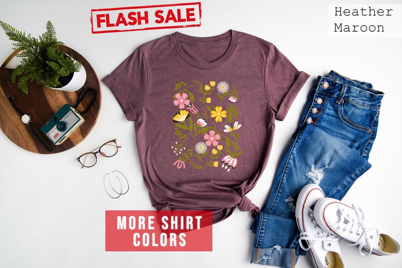 May include: A heather maroon t-shirt with a floral print featuring pink, yellow, and green flowers and butterflies. The shirt is displayed on a white background with a pair of jeans, white sneakers, and a wooden tray with a camera and glasses.
