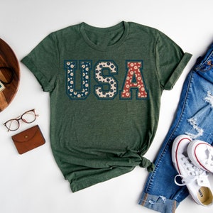 以下が含まれることがあります： フローラルデザインの「USA」の文字が入ったグリーンのTシャツ。文字は青と白、赤と白の花で埋められています。このシャツは、ブルージーンズと白いスニーカーと組み合わせています。