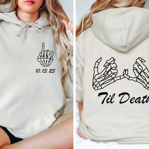May include: Cream-colored hoodie with black skeleton hand graphics. The front features a hand giving the middle finger and the date "10.13.23". The back displays two skeleton hands forming a promise with the text "Til Death".
