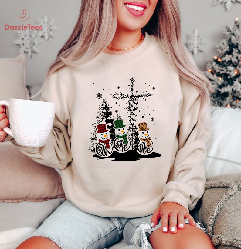 May include: Beige sweatshirt featuring a Christmas design with three snowmen wearing hats and scarves. The snowmen have the words "Faith, Hope, Love" below them. A cross and trees are also part of the design. The brand "DazzleTees" is printed on the top left.