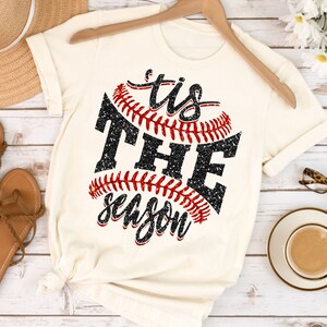 May include: A white t-shirt featuring a black and red baseball graphic with the text "'tis the season" in black glitter.