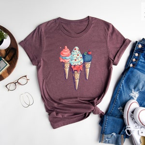 May include: A heather maroon t-shirt with a graphic of three ice cream cones. The ice cream cones are decorated with sprinkles, cherries, and a swirl of blue and pink ice cream.