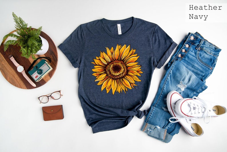 May include: A heather navy blue t-shirt with a yellow sunflower graphic. The shirt is laid flat on a white surface with a pair of jeans, a watch, a camera, glasses, and a wallet.