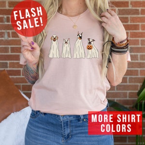 Op de afbeelding: Een lichtroze t-shirt met een grafische print van vier honden in witte spookkostuums. E&eacute;n hond houdt een kleine oranje pompoen vast. Het shirt bevat de tekst FLASH SALE en MORE SHIRT COLORS in rode kaders.
