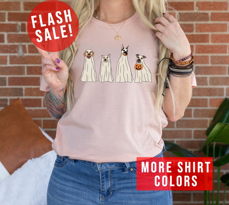 Op de afbeelding: Een lichtroze t-shirt met een grafische print van vier honden in witte spookkostuums. E&eacute;n hond houdt een kleine oranje pompoen vast. Het shirt bevat de tekst FLASH SALE en MORE SHIRT COLORS in rode kaders.